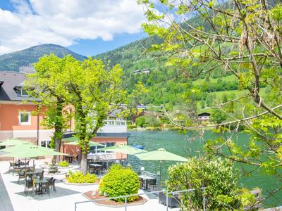 Terrasse mit Tischen und Stühlen am See, umgeben von grünen Bäumen und Berglandschaft.