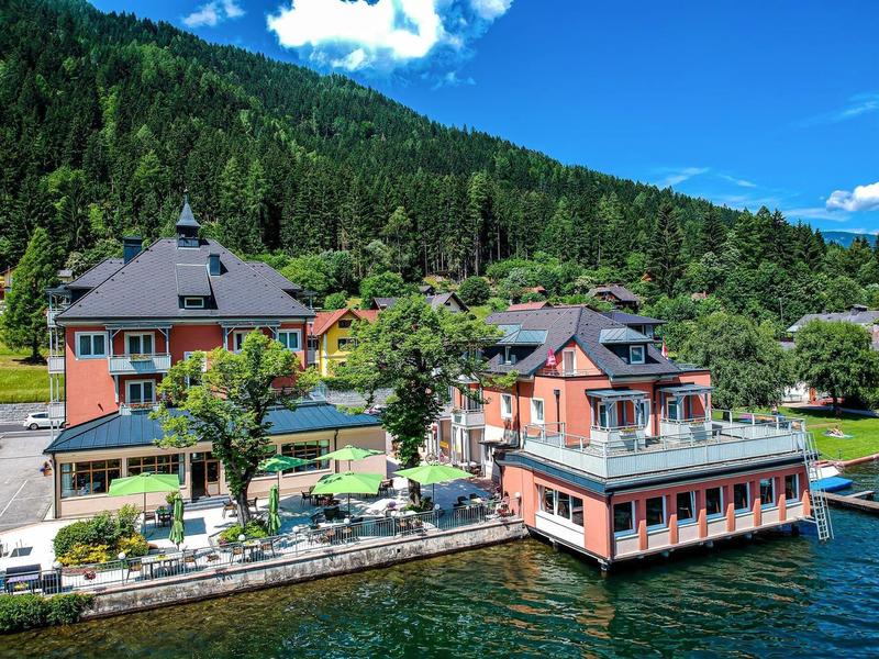 Uitzicht op een hotel aan een meer met een terras, omgeven door groene heuvels onder een blauwe lucht.