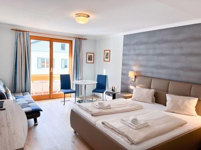 Bright hotel room with large bed, desk, chairs, and balcony with large windows.