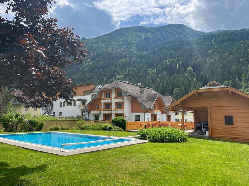 Green hotel grounds with outdoor pool, trees, and mountains in the background under cloudy sky.