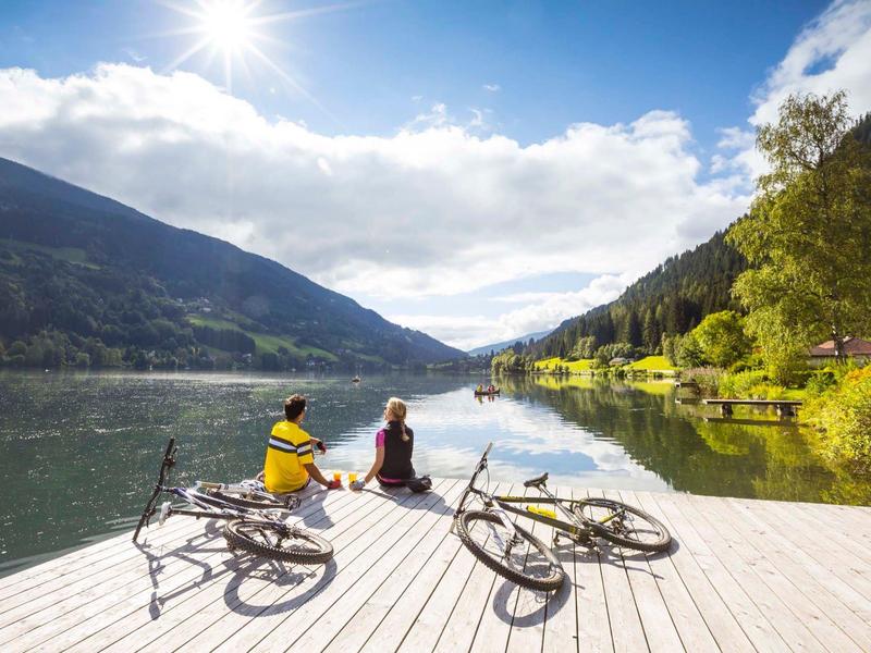 Two people sit on a lake dock with bikes, surrounded by mountains and sunlight.