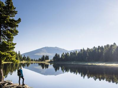 Ein Mann mit Rucksack steht am Ufer eines ruhigen Sees, umgeben von Bäumen und Bergen im Hintergrund.