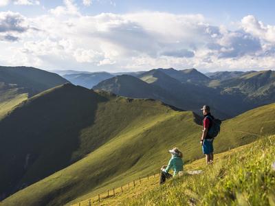 Zwei Wanderer auf grünem Berghang mit weiter Aussicht auf bewachsene Hügel und blauen Himmel.