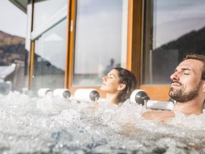 Zwei Personen entspannen in einem Whirlpool vor großen Fenstern mit Bergblick.