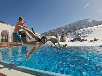 Zwei Junge springen spielerisch in einen Außenpool bei sonnigem, klarem Himmel und schneebedeckten Bergen.