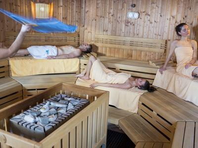 Menschen entspannen in einer Sauna mit Holzwänden und einem Ofen mit Steinen in der Mitte.