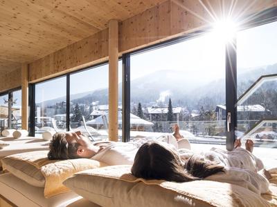 Zwei Personen entspannen in Holzhaus mit großen Fenstern und Ausblick auf verschneite Berge.