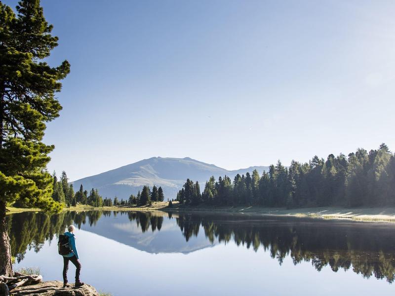 Ein Mann mit Rucksack steht am Ufer eines ruhigen Sees, umgeben von Bäumen und Bergen im Hintergrund.