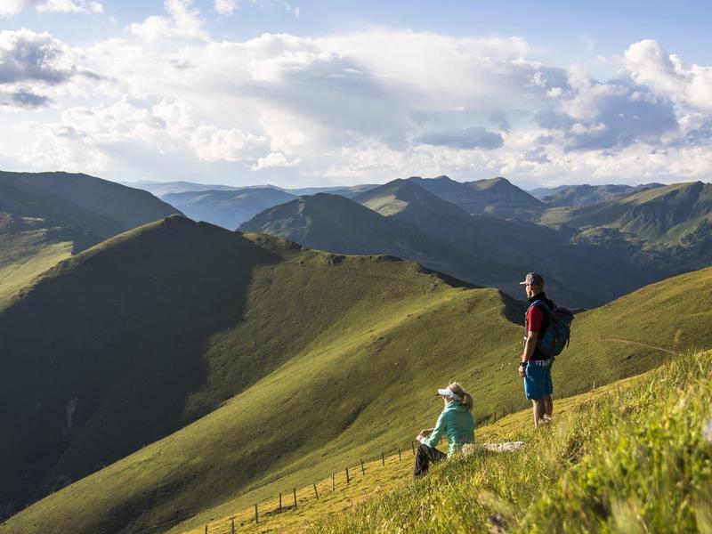 Zwei Wanderer auf grünem Berghang mit weiter Aussicht auf bewachsene Hügel und blauen Himmel.