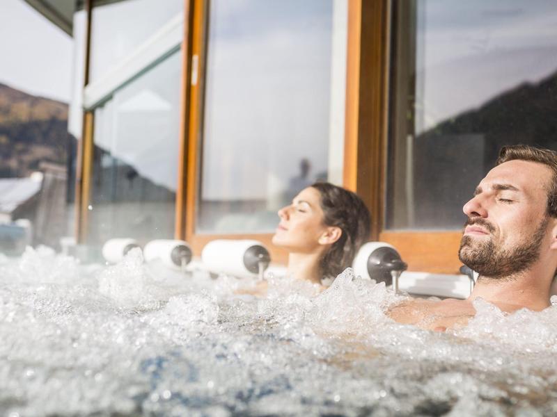 Zwei Personen entspannen in einem Whirlpool vor großen Fenstern mit Bergblick.