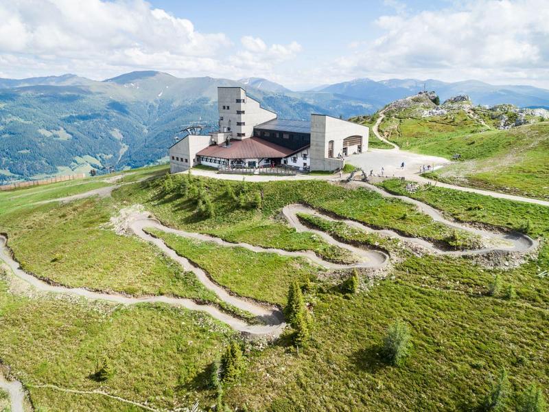 Grüne Berge mit gewundenen Wegen und einem Gebäude mit Aussichtsturm unter blauem Himmel.