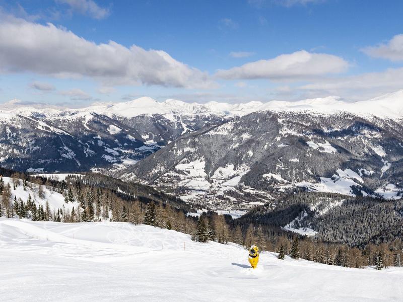 Winterlandschaft mit verschneiten Bergen und blauem Himmel, Skifahrer auf der Piste im Vordergrund.