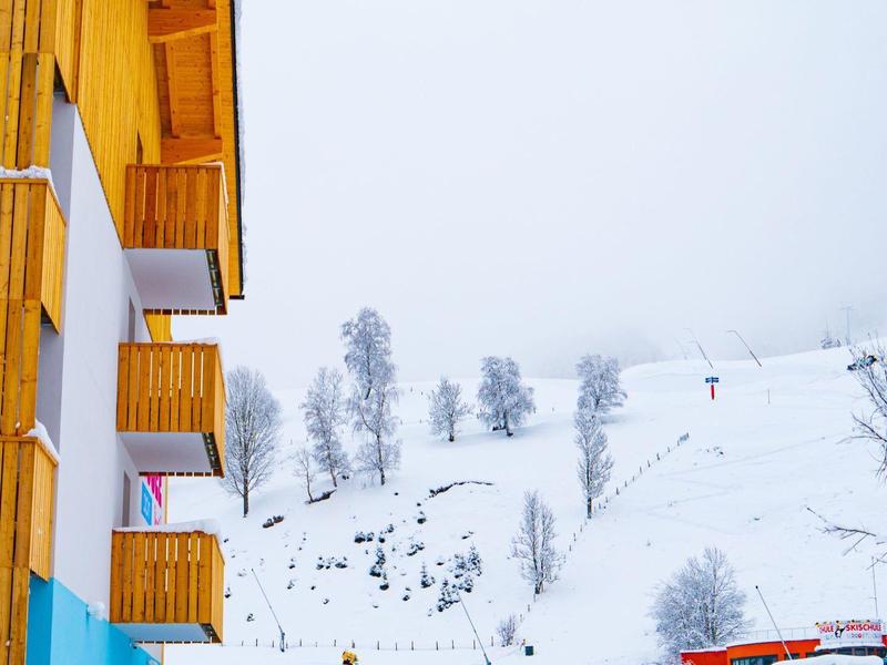 Holzchalets mit Balkonen vor schneebedecktem Berg und Skilift bei Nebel.