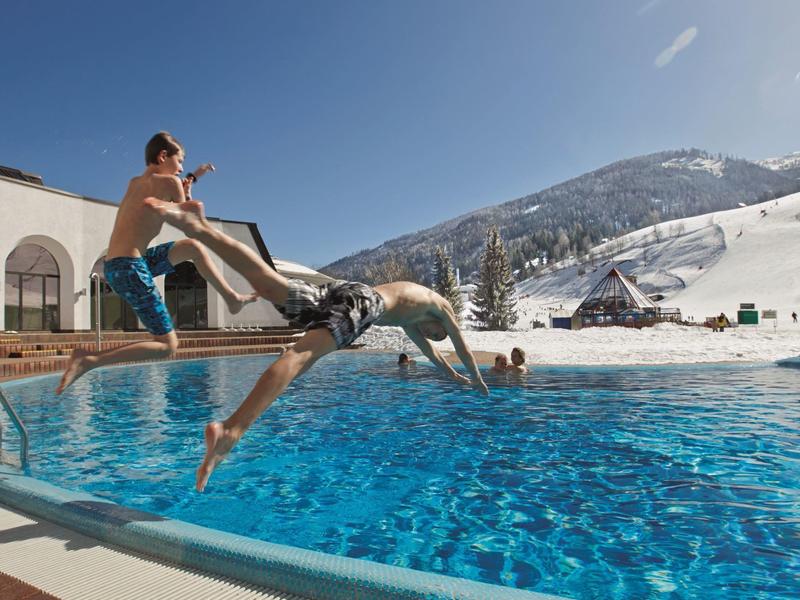 Zwei Junge springen spielerisch in einen Außenpool bei sonnigem, klarem Himmel und schneebedeckten Bergen.