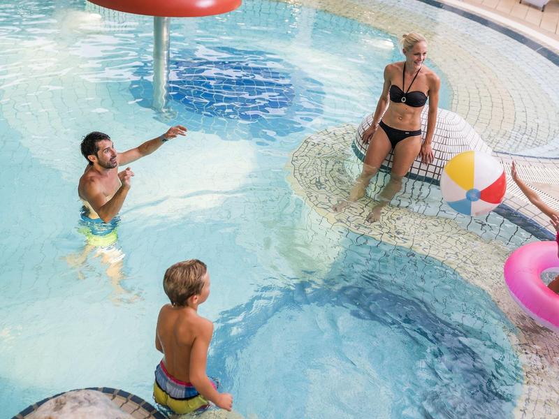 Eine Familie spielt im flachen Pool mit Wasserspilzen und buntem Ball, umgeben von Liegestühlen.