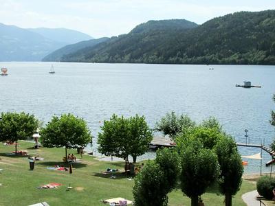 Grüne Parkanlage mit Bäumen vor einem ruhigen See und bewaldeten Hügeln im Hintergrund