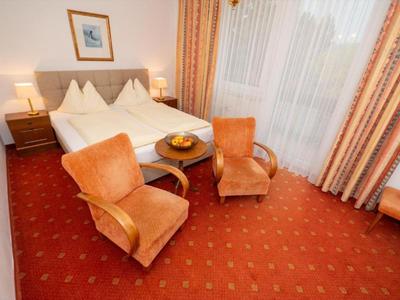 Gemütliches Hotelzimmer mit Doppelbett, zwei Sesseln und Tisch vor einem großen Fenster mit Gardinen.