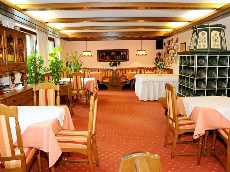 Gemütlicher Gastraum mit Holzmöbeln, karierten Sesseln, roten Teppich und grünem Kachelofen.