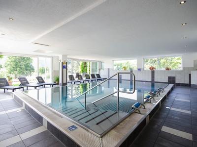 Helles Hallenbad mit großen Fenstern, Liegestühlen und einer Treppe zum Wasser.