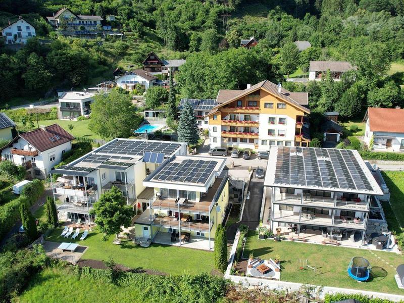 Mehrstöckige Wohnhäuser mit Solarzellen, umgeben von Grünflächen und Bäumen in ländlicher Umgebung.