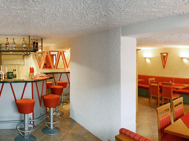 Innenraum einer Bar mit orangen Hockern, Tischen und Holzmöbeln, weiß gestrichene Wände, Decke strukturiert.