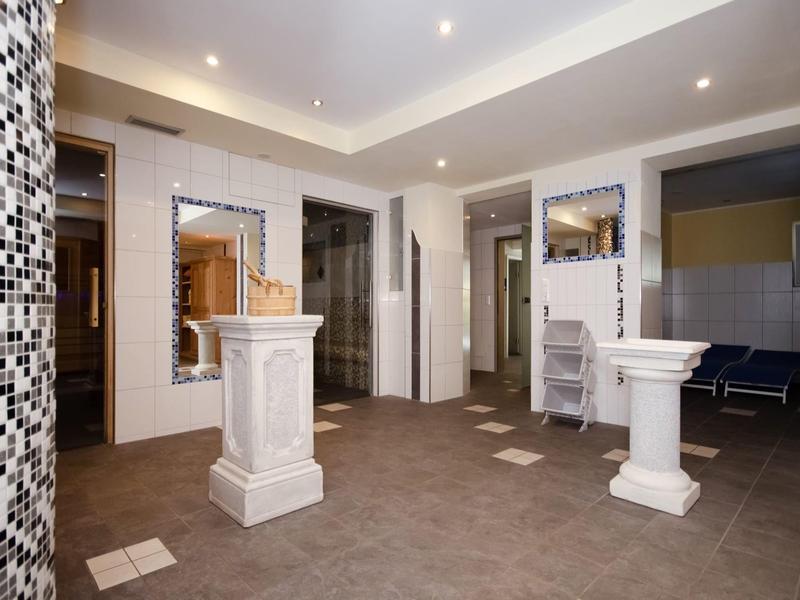 Moderner, heller Raum mit weißen Säulen, Mosaikwand, Spiegeln und offenen Türrahmen.