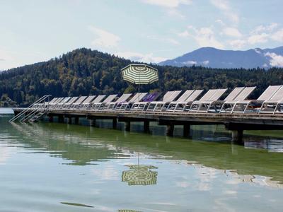 Langer Steg mit Sonnenliegen am ruhigen See vor bewaldeten Hügeln und Bergen.