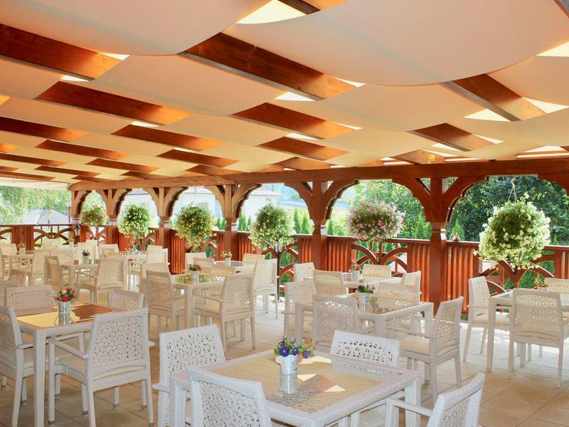 Überdachte Terrasse eines Restaurants mit weißen Tischen und Stühlen und Blick auf grüne Bäume.