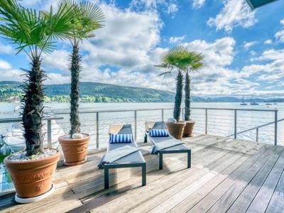 Terrasse mit Liegestühlen, Palmen und Blick auf einen See unter bewölktem Himmel