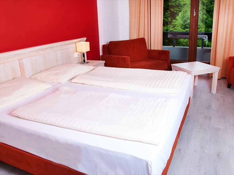 Hotelzimmer mit großem Doppelbett, roten Sesseln und hellen Vorhängen neben Fenster