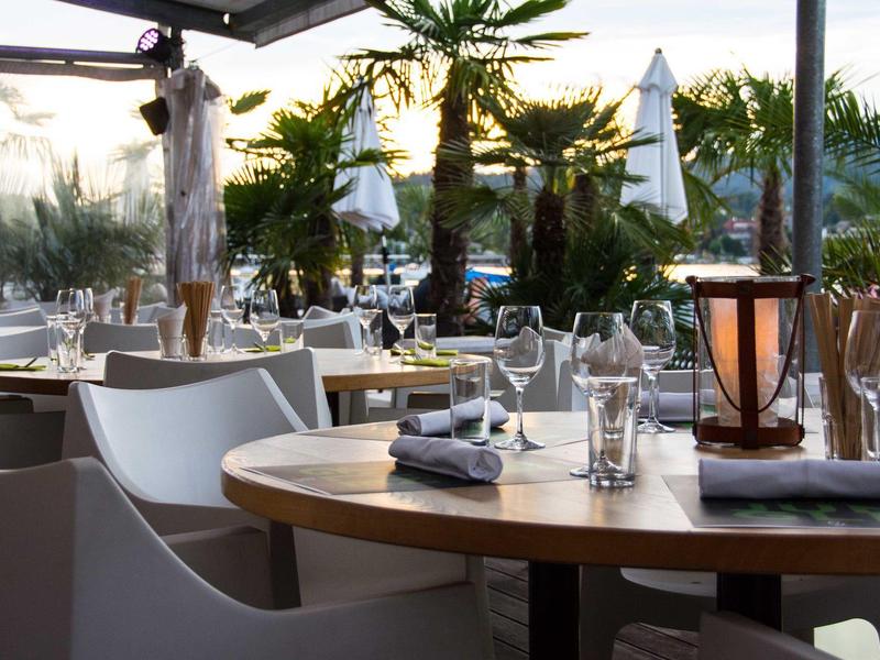 Elegantes Restaurantterrasse mit modernen weißen Stühlen, gedeckten Tischen und Palmen bei Sonnenuntergang.