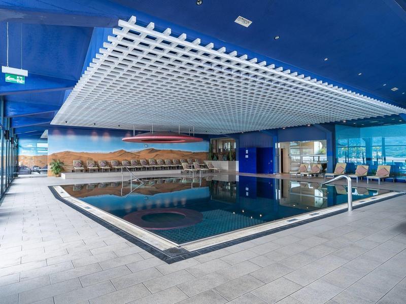 Moderner Innenpool mit blauer Decke, großen Fenstern und Entspannungsbereichen.