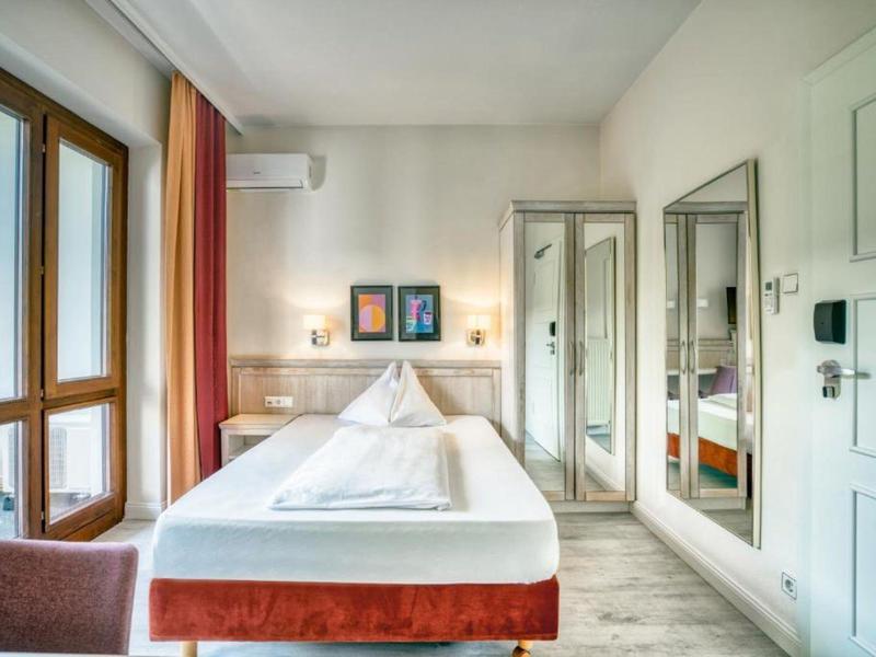 Hell eingerichtetes Hotelzimmer mit großem Bett, Spiegelkleiderschrank und Blick auf Balkon.