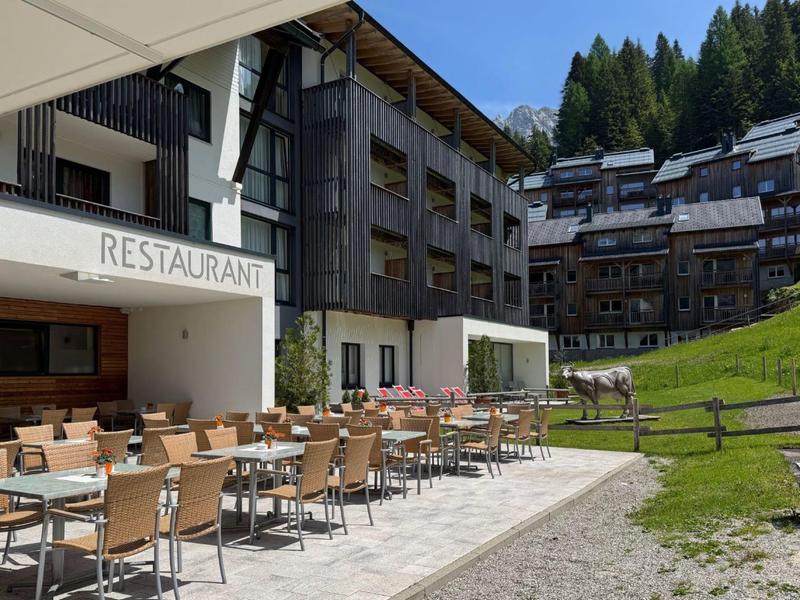 Buitenrestaurant met houten tafels en stoelen naast een meerlaagse hotel, gelegen in berglandschap.