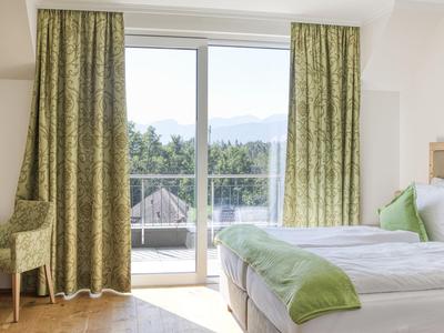 Helles Hotelzimmer mit Doppelbett, grün gemusterten Vorhängen und Balkon mit Aussicht auf Bäume.