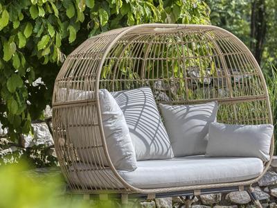 Ein rundes Rattansofa mit hellen Kissen steht im schattigen Garten unter einem Baum.