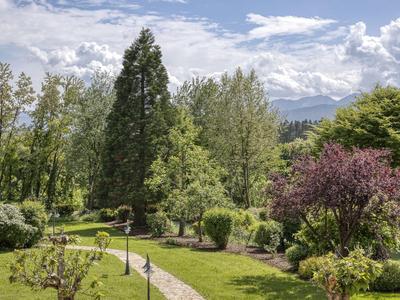 Grüner Garten mit gepflastertem Weg, Bäumen und blühenden Sträuchern unter bewölktem Himmel.