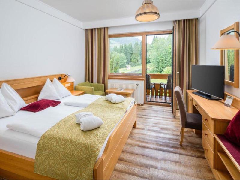 Bright hotel room with double bed, balcony, and view of green trees.