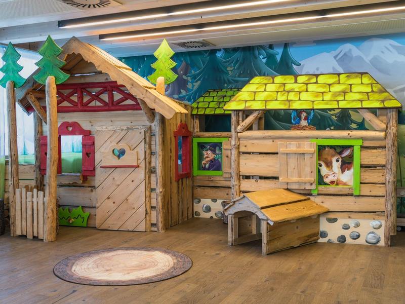 Aire de jeux en bois intérieur avec petites cabanes, toit coloré et décorations d'arbres.