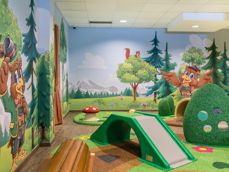 Espace de jeu intérieur coloré avec des fresques forestières, des structures d'escalade et des équipements de jeu verts doux.