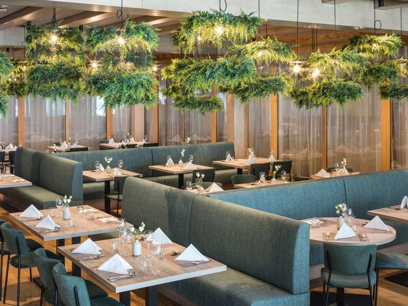 Restaurant moderne avec banquettes vertes, tables en bois et plantes suspendues avec lumières.