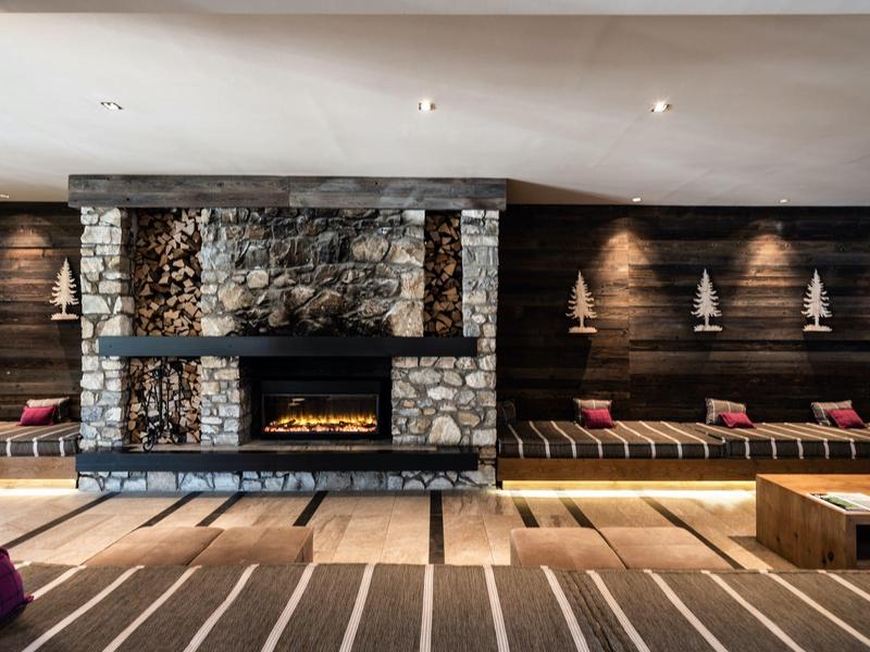 Espace lounge confortable avec cheminée en pierre, murs en bois et sièges avec coussins rouges.