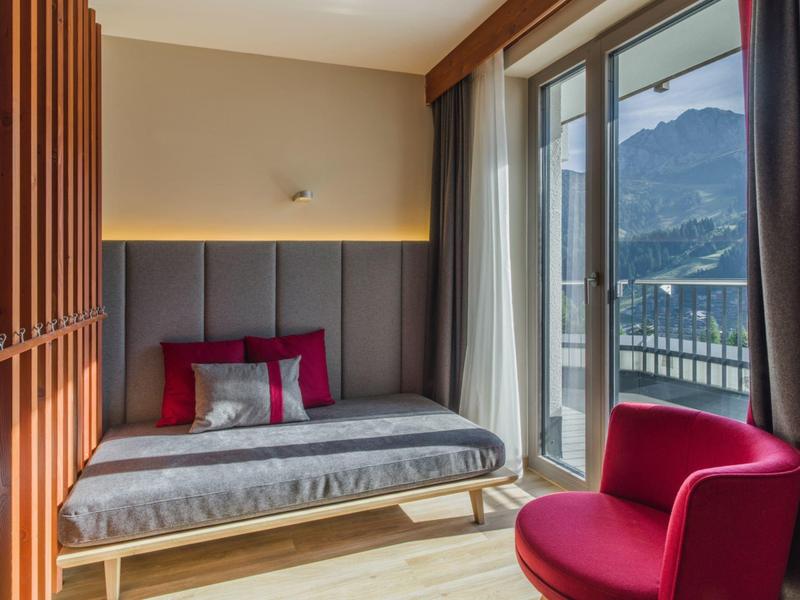 Chambre d'hôtel moderne avec lit, fauteuil rouge et balcon offrant vue sur les montagnes.