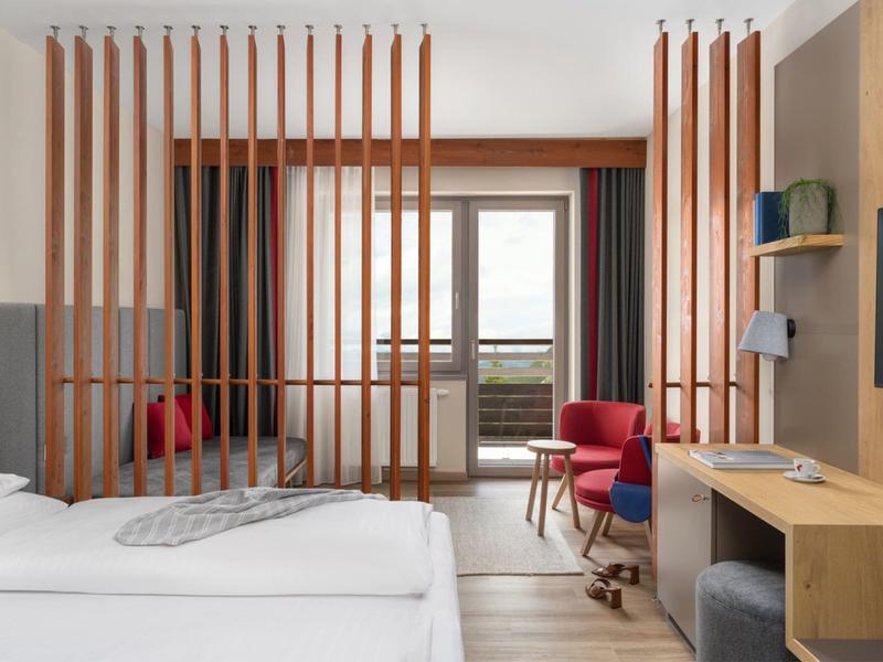 Chambre d'hôtel avec deux lits, cloisons en bois, fauteuils rouges et porte balcon avec vue extérieure.