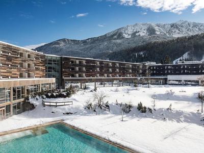 Modernes Hotel mit Außenpool und schneebedecktem Gelände vor Bergen bei blauem Himmel.