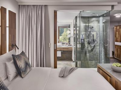 Modernes Hotelzimmer mit Bett, Kissen, Glasdusche und Holzmöbeln in neutralen Tönen.