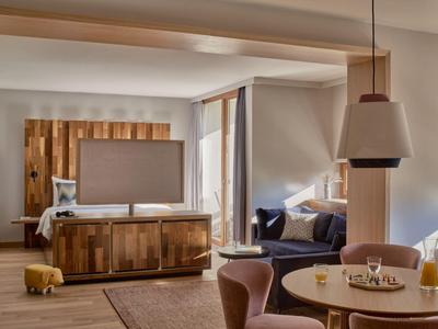 Gemütliches Hotelzimmer mit Holzmöbeln, Sofa, Fernseher und großem Fenster mit Vorhängen.