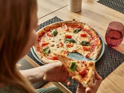 Person hält ein Stück Pizza mit Basilikumblättern an einem holzfarbenen Tisch mit rotem Glas.