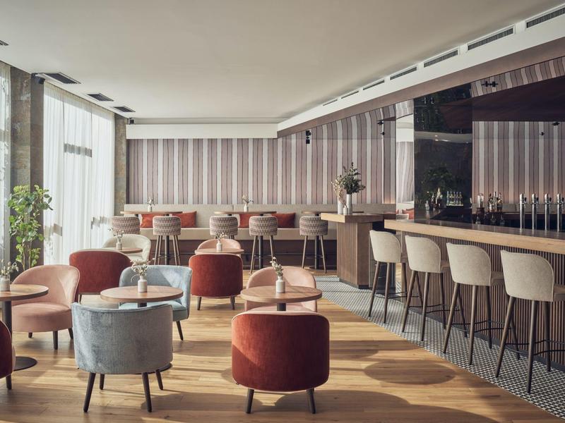 Nowoczesny bar hotelowy z tapicerowanymi fotelami, stołkami barowymi i dużymi oknami wpuszczającymi światło dzienne.