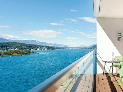 Balkon mit Glasgeländer, grünem Stuhl und Tisch, blickt auf blauen See und bewaldete Berge.